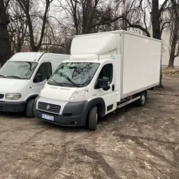 Dwa białe samochody dostawcze, Opel Movano i Fiat Ducato z zabudową kontenerową, stoją obok siebie na błotnistym placu miejskim z widocznymi śladami opon.