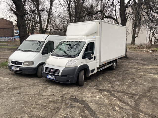 Dwa białe samochody dostawcze, Opel Movano i Fiat Ducato z zabudową kontenerową, stoją obok siebie na błotnistym placu miejskim z widocznymi śladami opon.