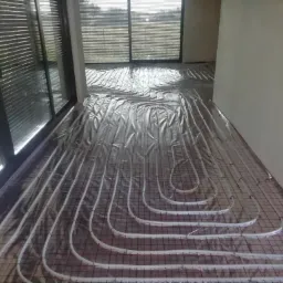 Białe rury ogrzewania podłogowego ułożone na srebrnej folii izolacyjnej z czerwoną siatką w jasnym pokoju z dużymi oknami.