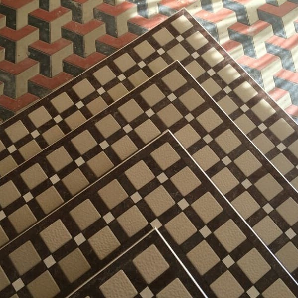 Ułożona pod kątem mozaika podłogowa z płytek w geometryczne wzory: szare kwadraty z ciemnobrązowymi obwódkami i małymi białymi akcentami, w tle trójwymiarowy wzór w odcieniach czerwieni i szarości.