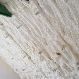 Poddasze z drewnianymi belkami pokryte grubą warstwą białej pianki izolacyjnej, zabezpieczonej metalowymi klamrami. W tle widoczny fragment ściany z cegły i okno.