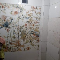 PAW-exBUD Prace Remontowo-Wykończeniowe - Fragment ściany w trakcie układania płytek z motywem kwiatowym i papugami, widoczne krzyżyki dystansowe, gładkie, białe płytki na sąsiedniej ścianie, otwór rewizyjny w górnej części ściany.