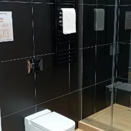 Nowoczesna łazienka z czarnymi płytkami, wiszącą toaletą, grzejnikiem drabinkowym z białym ręcznikiem i kabiną prysznicową ze szklanymi drzwiami.