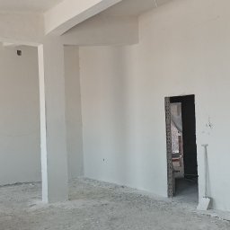 PAW-exBUD Prace Remontowo-Wykończeniowe - Wnętrze w stanie deweloperskim po wykonaniu tynków: surowe, białe ściany z widocznymi przewodami elektrycznymi zwisającymi z sufitu, betonowa podłoga z resztkami zaprawy i narzędzia w przejściu...