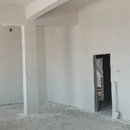 Wnętrze w stanie deweloperskim po wykonaniu tynków: surowe, białe ściany z widocznymi przewodami elektrycznymi zwisającymi z sufitu, betonowa podłoga z resztkami zaprawy i narzędzia w przejściu...