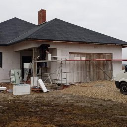 PAW-exBUD Prace Remontowo-Wykończeniowe - Budowa domu jednorodzinnego: murarz na rusztowaniu układa bloczki z betonu komórkowego, widoczny fragment dachu pokrytego dachówką, biały samochód dostawczy w tle.