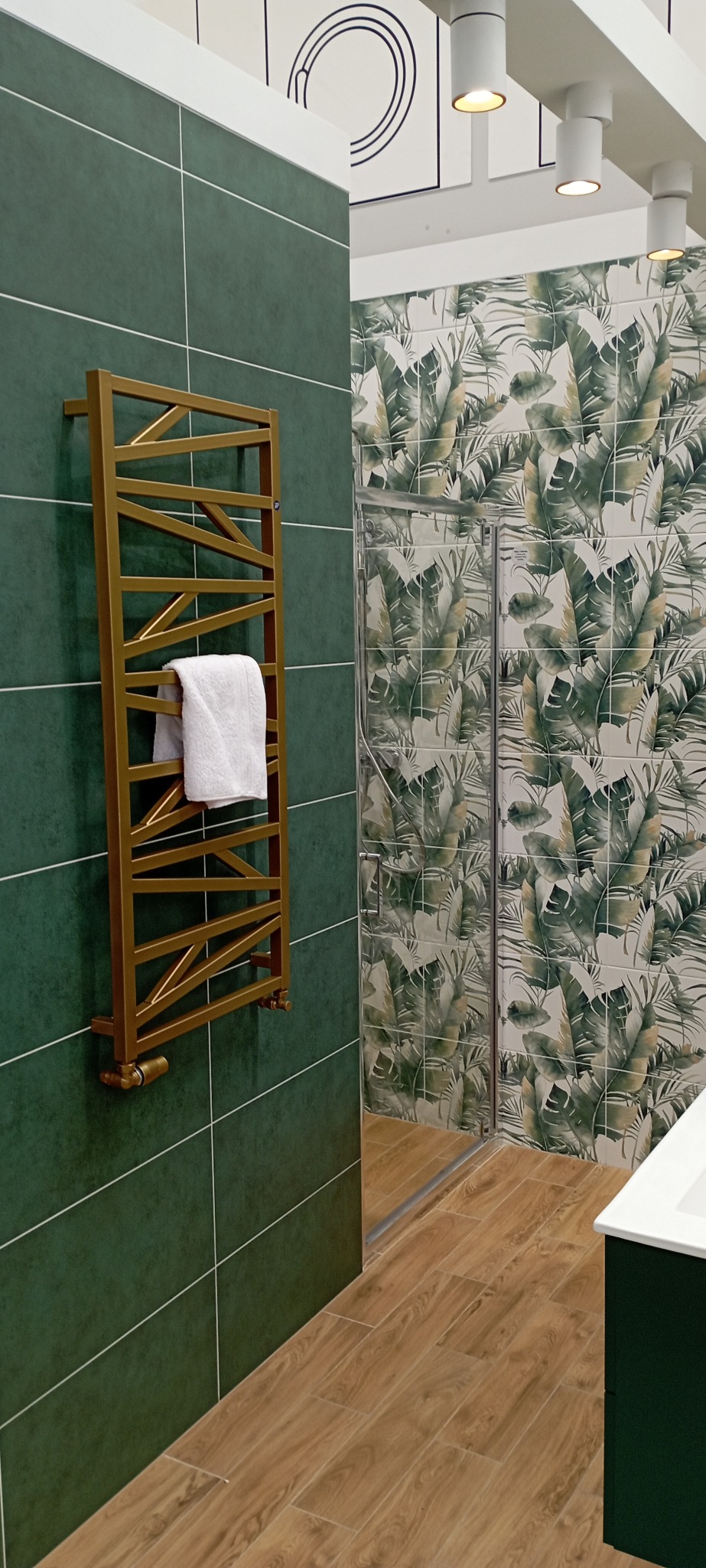 Nowoczesna łazienka z zielonymi płytkami, złotym grzejnikiem drabinkowym z ręcznikiem, podłogą imitującą drewno i kabiną prysznicową z motywem liści.