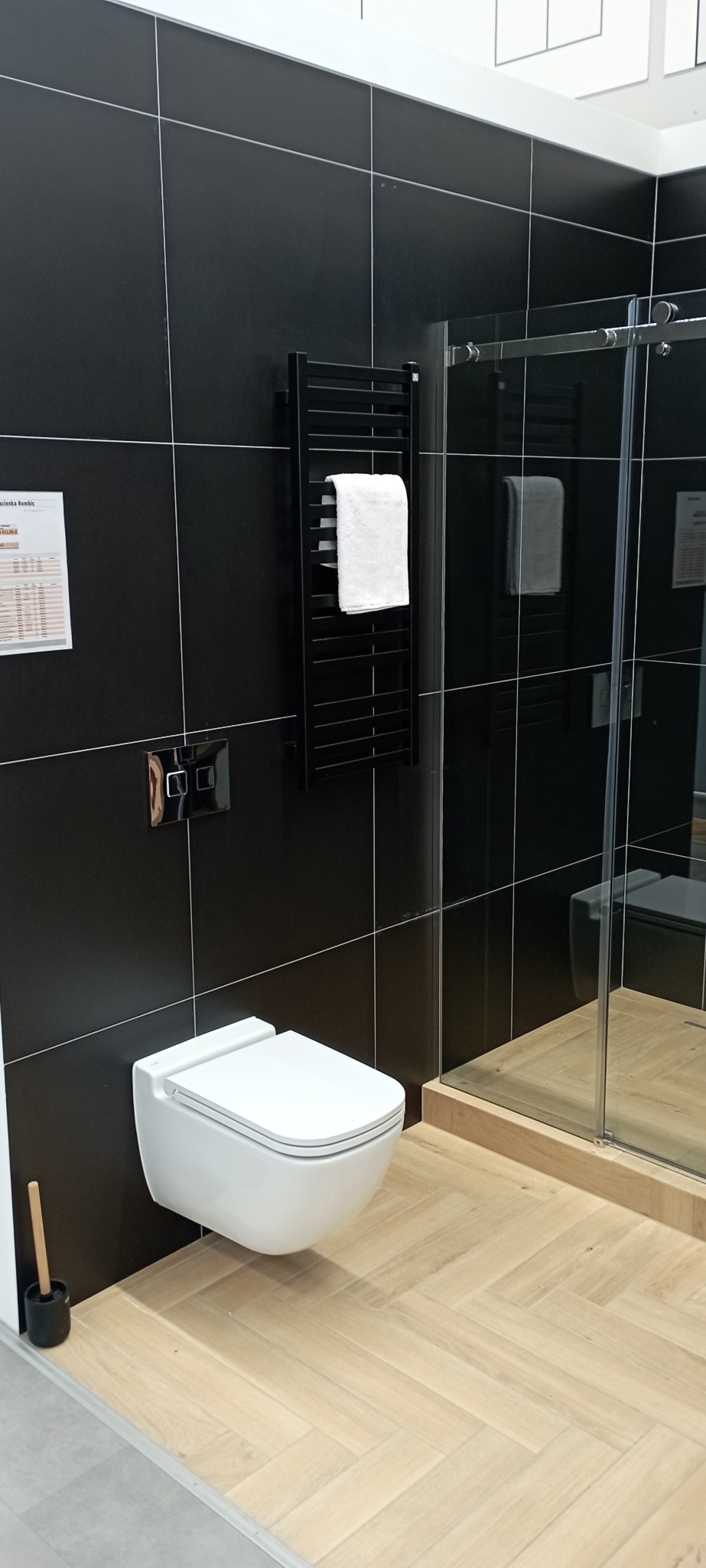 Nowoczesna łazienka z czarnymi płytkami, wiszącą toaletą, grzejnikiem drabinkowym z białym ręcznikiem i kabiną prysznicową ze szklanymi drzwiami.