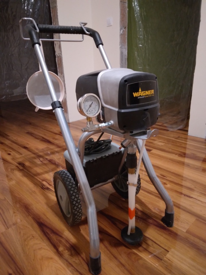 Agregat malarski Wagner PowerPainter 90 na kółkach, z manometrem i filtrem, stojący na parkiecie w pomieszczeniu przygotowanym do malowania, z folią ochronną w tle.