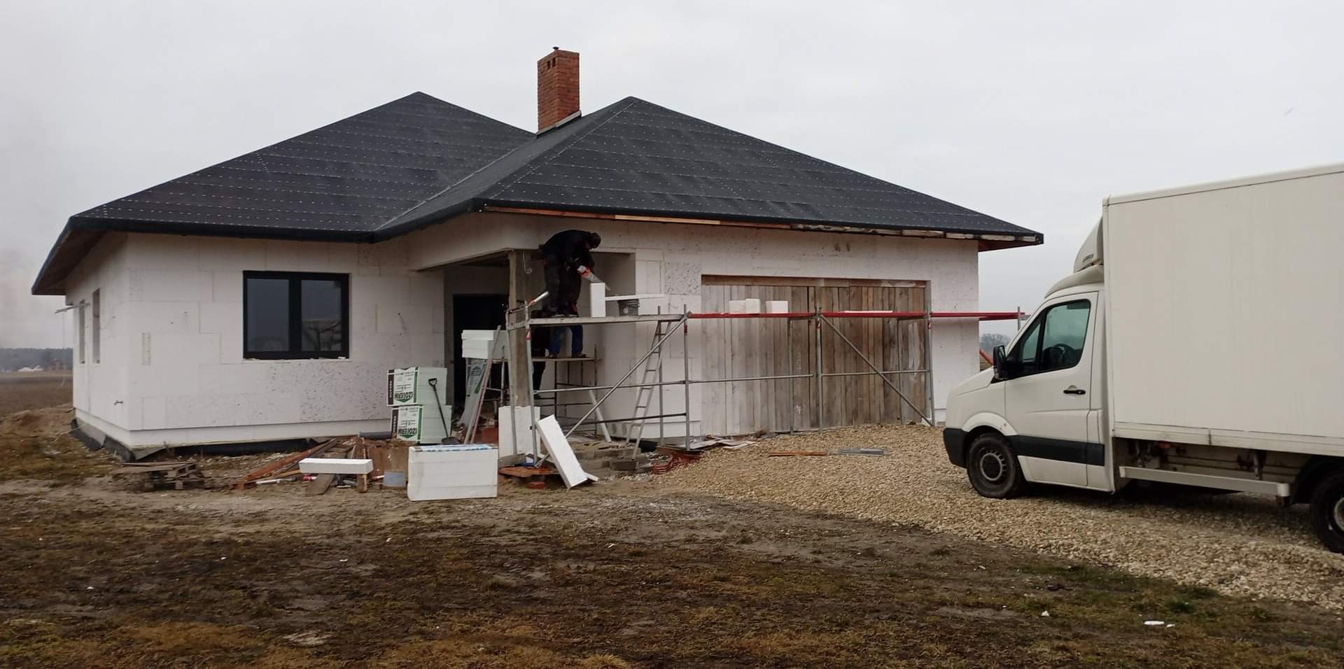 Budowa domu jednorodzinnego: murarz na rusztowaniu układa bloczki z betonu komórkowego, widoczny fragment dachu pokrytego dachówką, biały samochód dostawczy w tle.