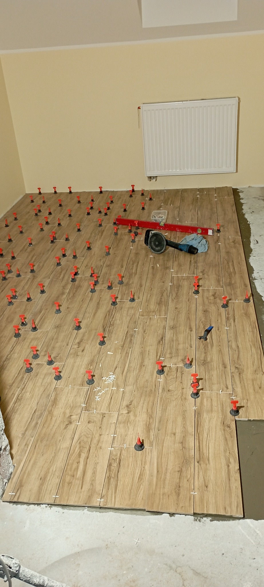 Układanie płytek podłogowych imitujących drewno z użyciem systemu poziomowania płytek, widoczna poziomica, szlifierka kątowa i fragment ściany z grzejnikiem.
