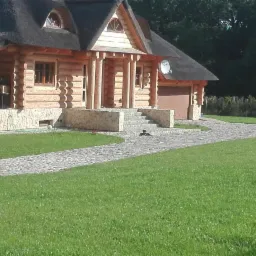 Drewniany dom z krytym strzechą dachem, kamienną podmurówką i ścieżką z kostki granitowej prowadzącą przez trawnik.