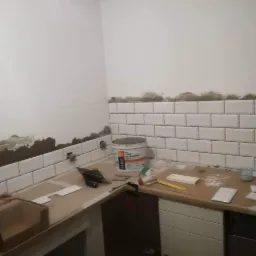 Ściana w trakcie układania białych, prostokątnych płytek, z widocznymi resztkami kleju i narzędziami glazurniczymi na blacie.