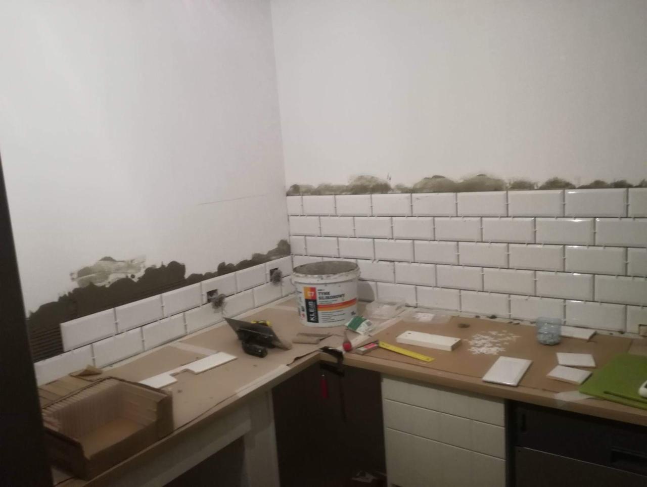 Ściana w trakcie układania białych, prostokątnych płytek, z widocznymi resztkami kleju i narzędziami glazurniczymi na blacie.