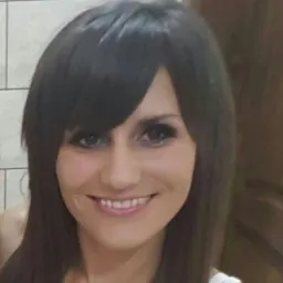 Portret uśmiechniętej kobiety z ciemnymi włosami, prawdopodobnie nauczycielki, z widocznym fragmentem białej bluzki i tłem z jasnych płytek oraz elementów drewnianych.