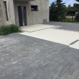 Ułożona kostka brukowa w dwóch odcieniach szarości i bieli, tworząca wzór przed domem z surową elewacją i ozdobną trawą.