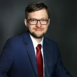 Portret mężczyzny w okularach, niebieskim garniturze i czerwonym krawacie na ciemnoszarym tle, z delikatnym uśmiechem.