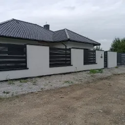 Nowoczesne ogrodzenie posesji z białymi słupkami i czarnymi, poziomymi panelami, na tle domu z ciemną dachówką, otoczone żwirem.