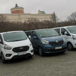 Rząd zaparkowanych bus&oacute;w r&oacute;żnych kolor&oacute;w (szary, biały, niebieski) na tle zamku w Lublinie, każdy z widoczną tablicą rejestracyjną.