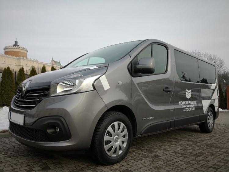 Szary van Renault z logo firmy 'New Car Prestige' na boku, zaparkowany na brukowanym placu z budynkiem w tle.