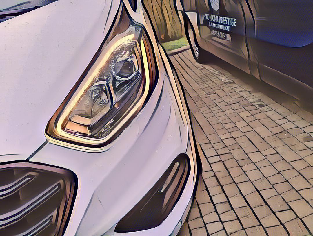 Zbliżenie na przedni reflektor białego samochodu z widocznym logo firmy 'Newcar Prestige' na ciemnym samochodzie w tle, na brukowanym podjeździe.