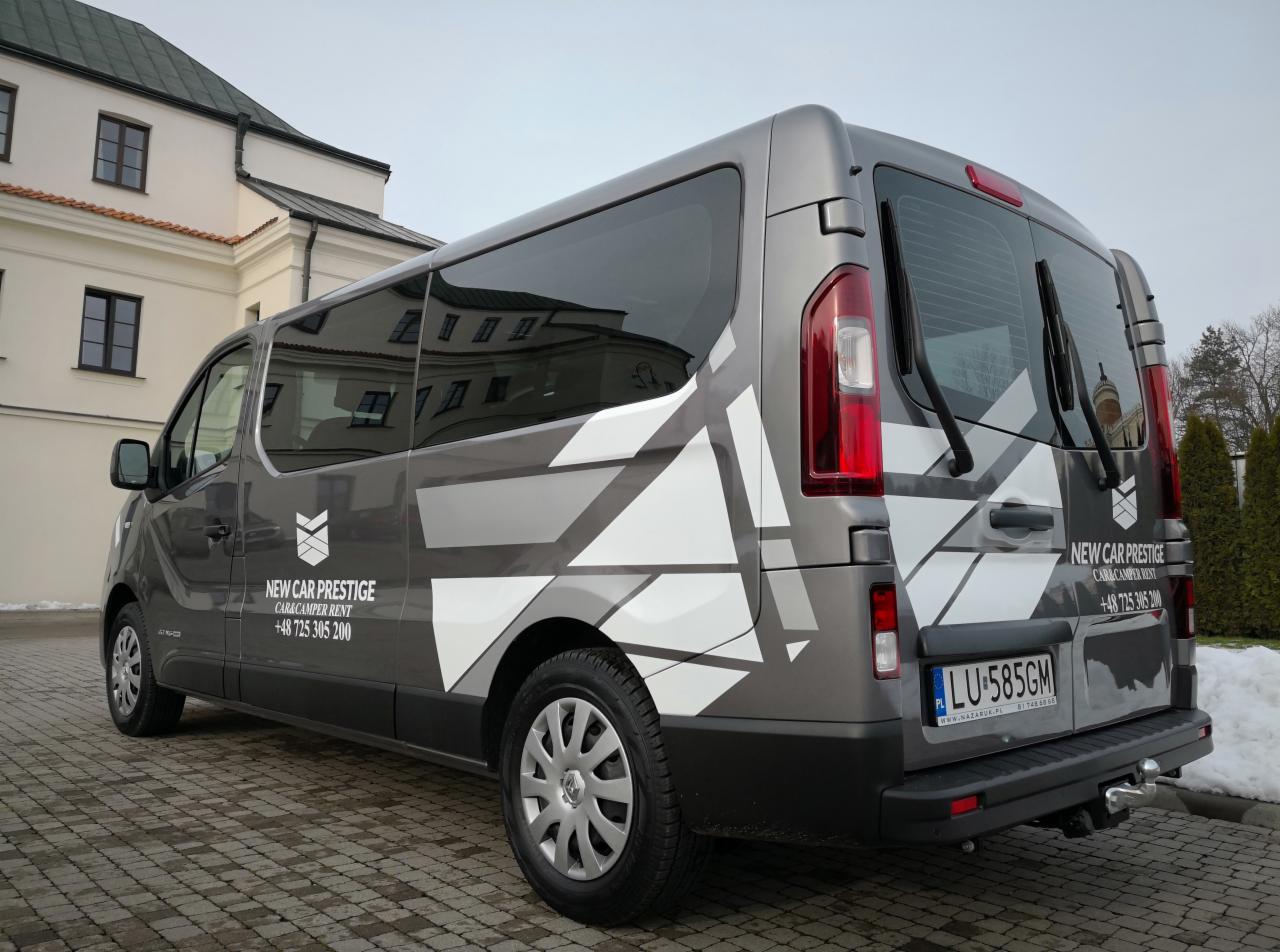 Szary van z logo i numerem telefonu wypożyczalni samochodów New Car Prestige, zaparkowany na brukowanej powierzchni, z budynkiem w tle.