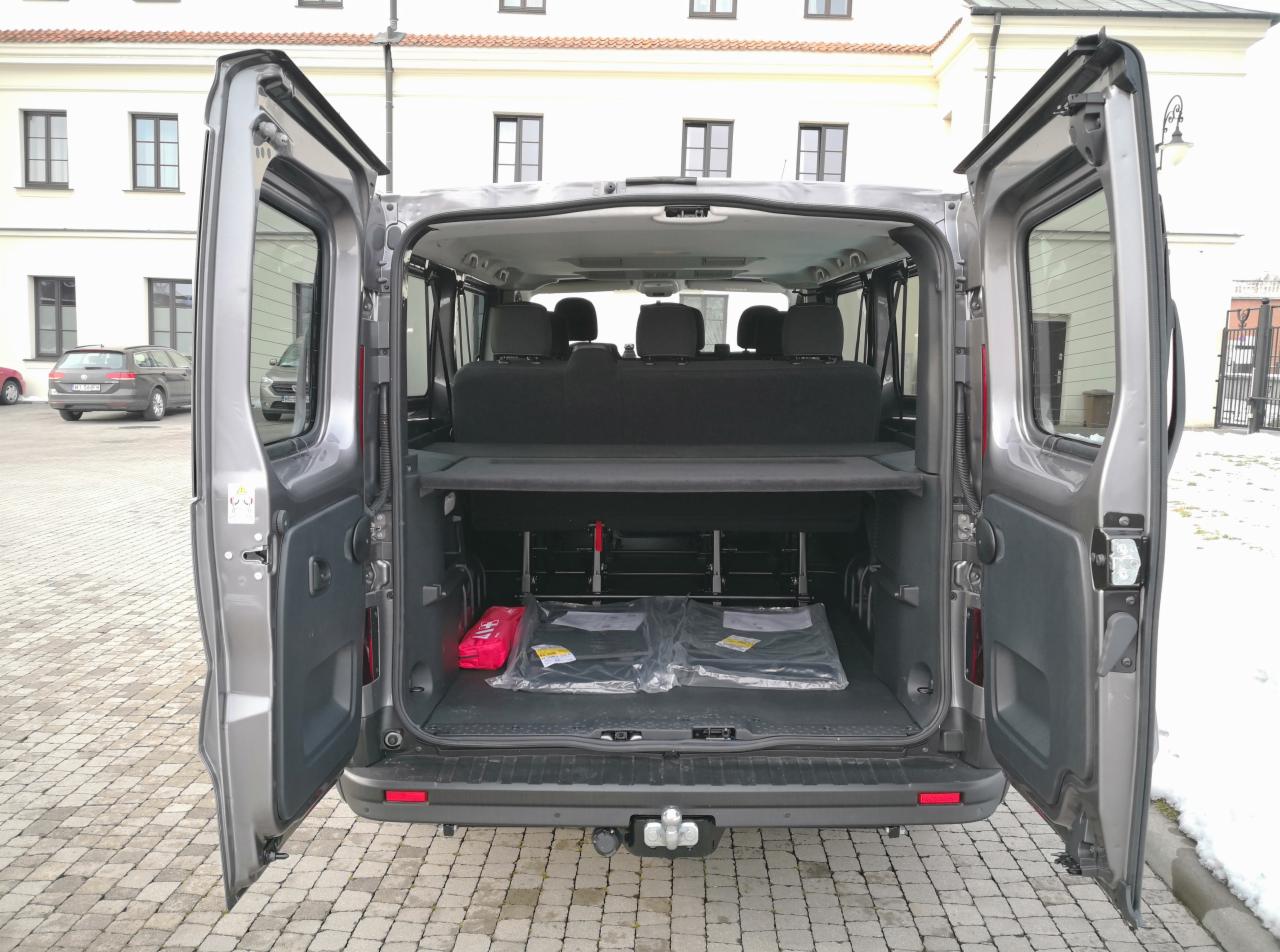 Otwarty bagażnik szarego vana z widoczną apteczką, nowymi dywanikami w folii i zabezpieczeniem przestrzeni bagażowej, na zewnątrz budynek z oknami.