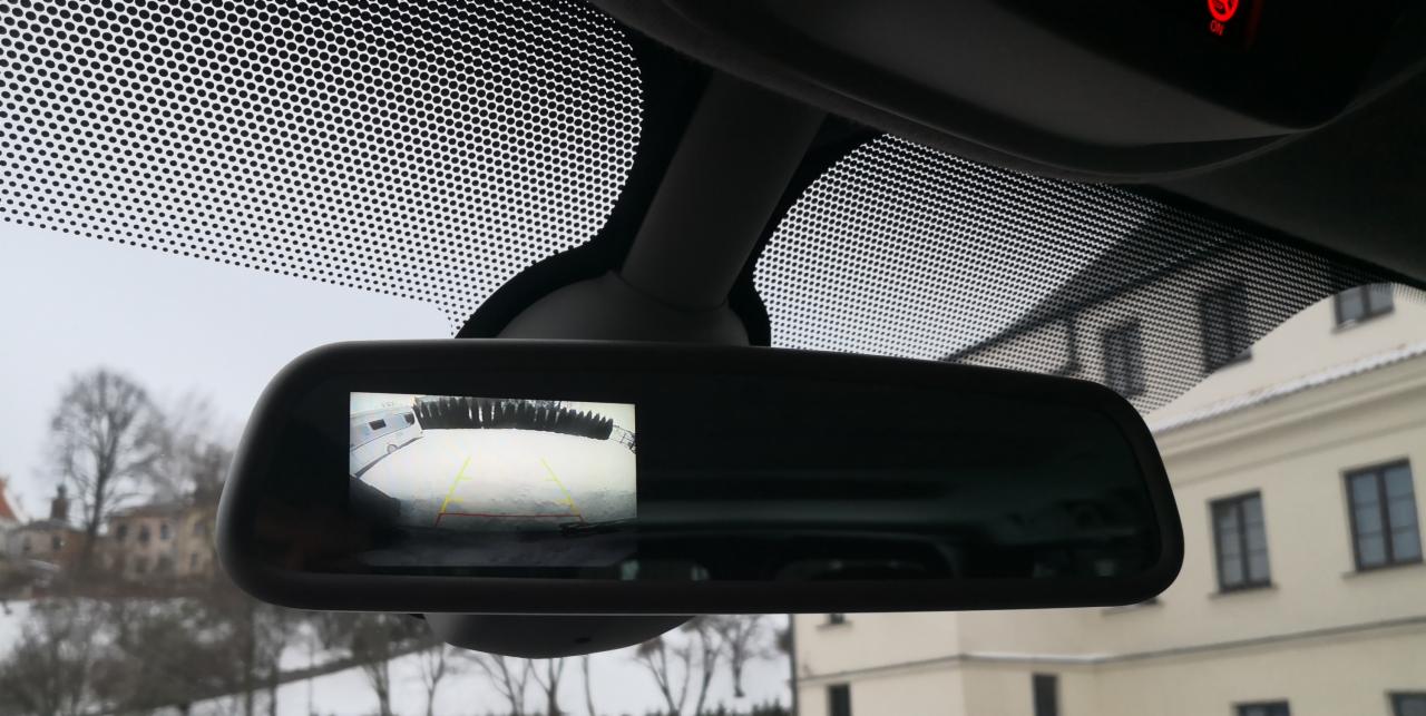 Wnętrze samochodu z widokiem na lusterko wsteczne, w którym wyświetlany jest obraz z kamery cofania, na zewnątrz widoczny budynek i drzewa w zimowej scenerii.