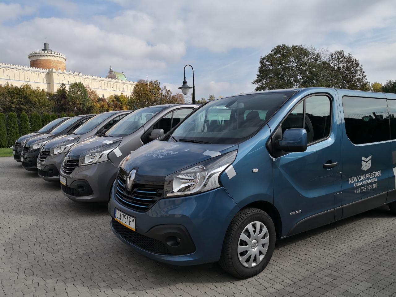Szereg zaparkowanych, nowych, niebieskich i szarych vanów Renault Trafic z logo firmy i numerem telefonu na karoserii, na tle brukowanego placu i budynku z wieżą.