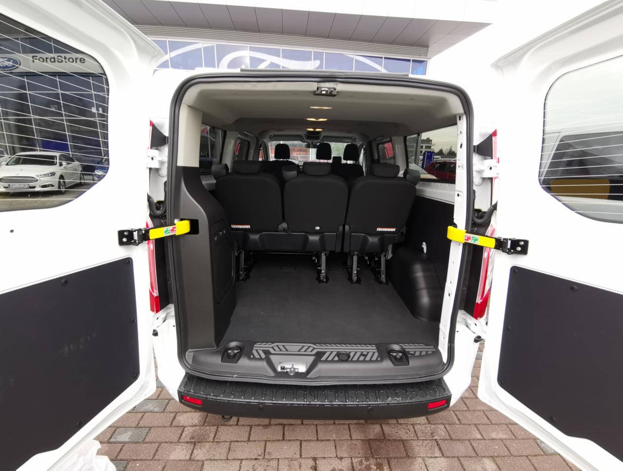 Wnętrze białego busa z otwartymi tylnymi drzwiami, widoczne trzy rzędy siedzeń dla pasażerów, zaparkowany przed salonem Forda.