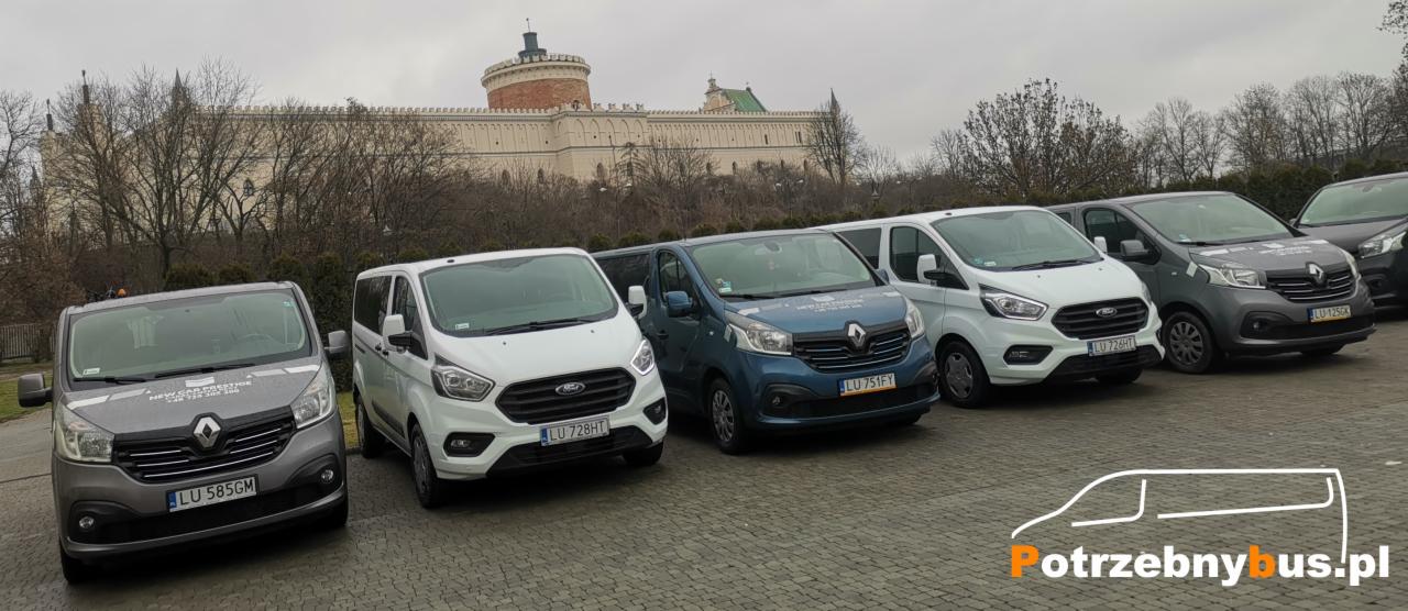 Rząd zaparkowanych busów różnych kolorów (szary, biały, niebieski) na tle zamku w Lublinie, każdy z widoczną tablicą rejestracyjną.