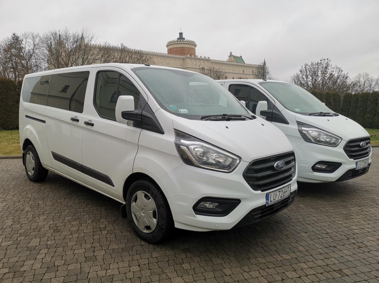 Dwa białe busy Ford Transit Custom zaparkowane na brukowanym placu przed budynkiem z wieżą w tle, widoczne tablice rejestracyjne LU 726HT i LU 728HT.