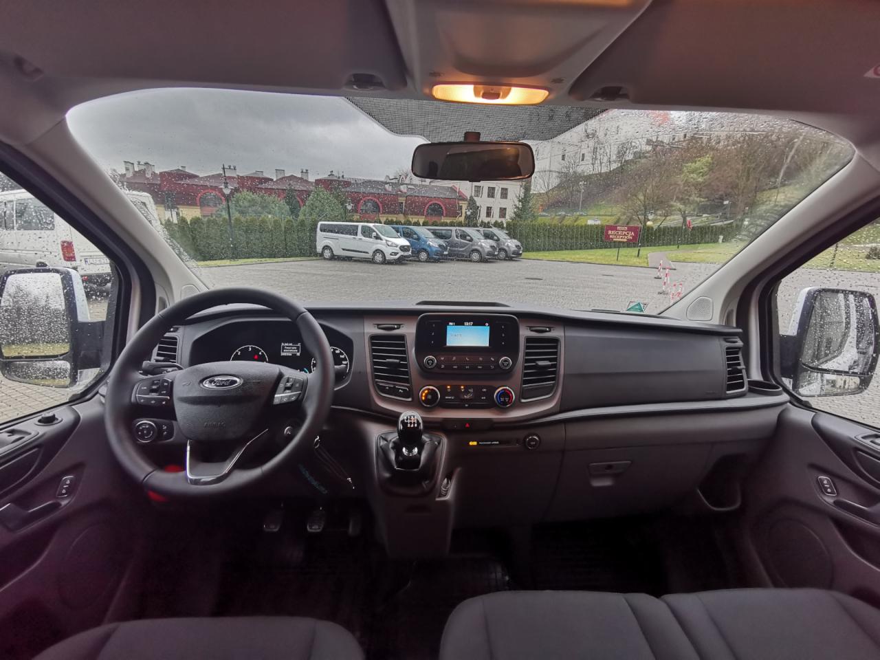 Wnętrze Forda Transita z manualną skrzynią biegów, widok z kabiny kierowcy na parking z kilkoma vanami w pochmurny dzień.