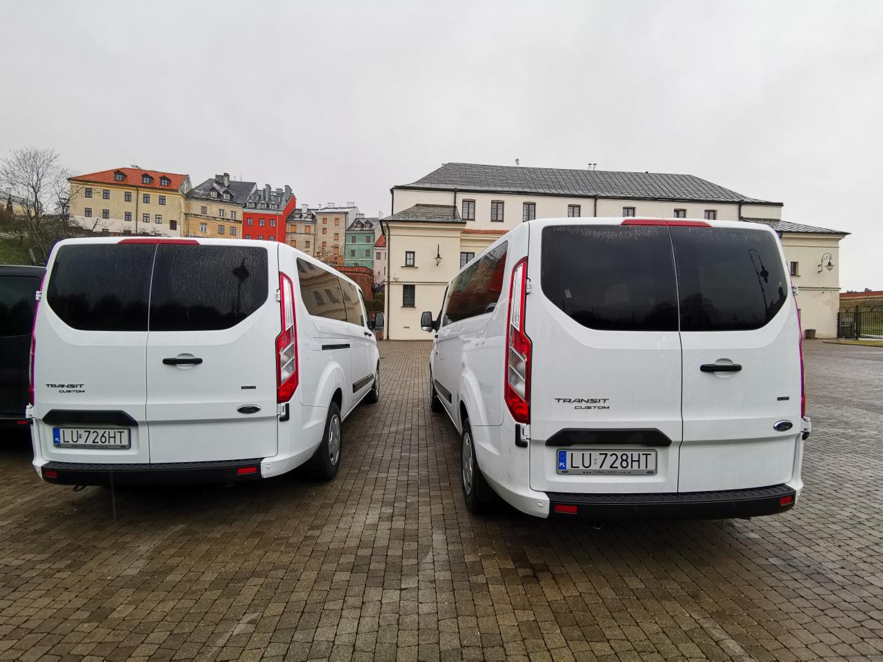 Dwa białe busy Ford Transit Custom zaparkowane tyłem obok siebie na brukowanym placu, w tle budynki o zróżnicowanej architekturze i kolorystyce.