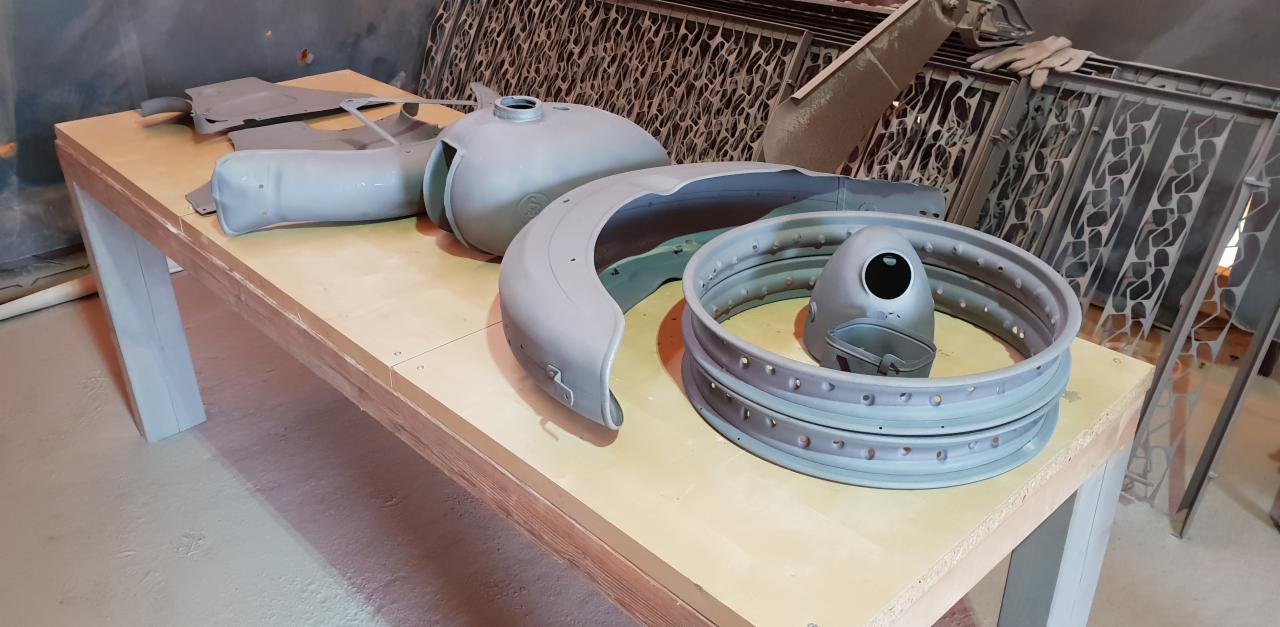 Elementy motocykla, w tym błotnik, zbiornik paliwa i obręcze kół, przygotowane do malowania proszkowego, leżące na stole w warsztacie.