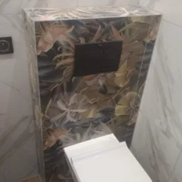 Podtynkowa toaleta Roca z czarnym przyciskiem spłukującym, obudowana płytkami w motywie tropikalnych liści, na tle marmurowych płytek ściennych.