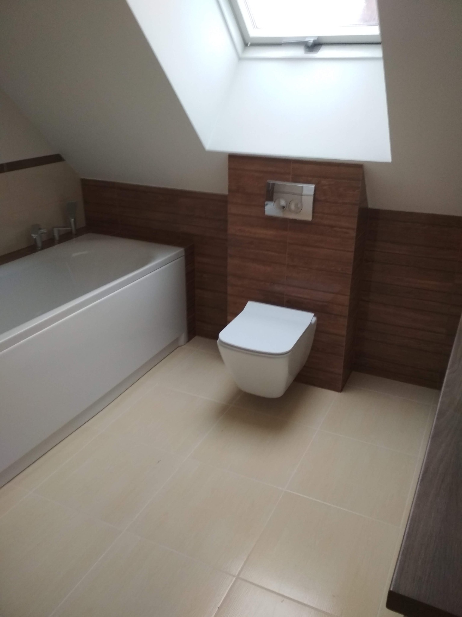 Nowoczesna łazienka na poddaszu z oknem dachowym, białą wanną i podwieszaną toaletą z brązowymi płytkami imitującymi drewno.