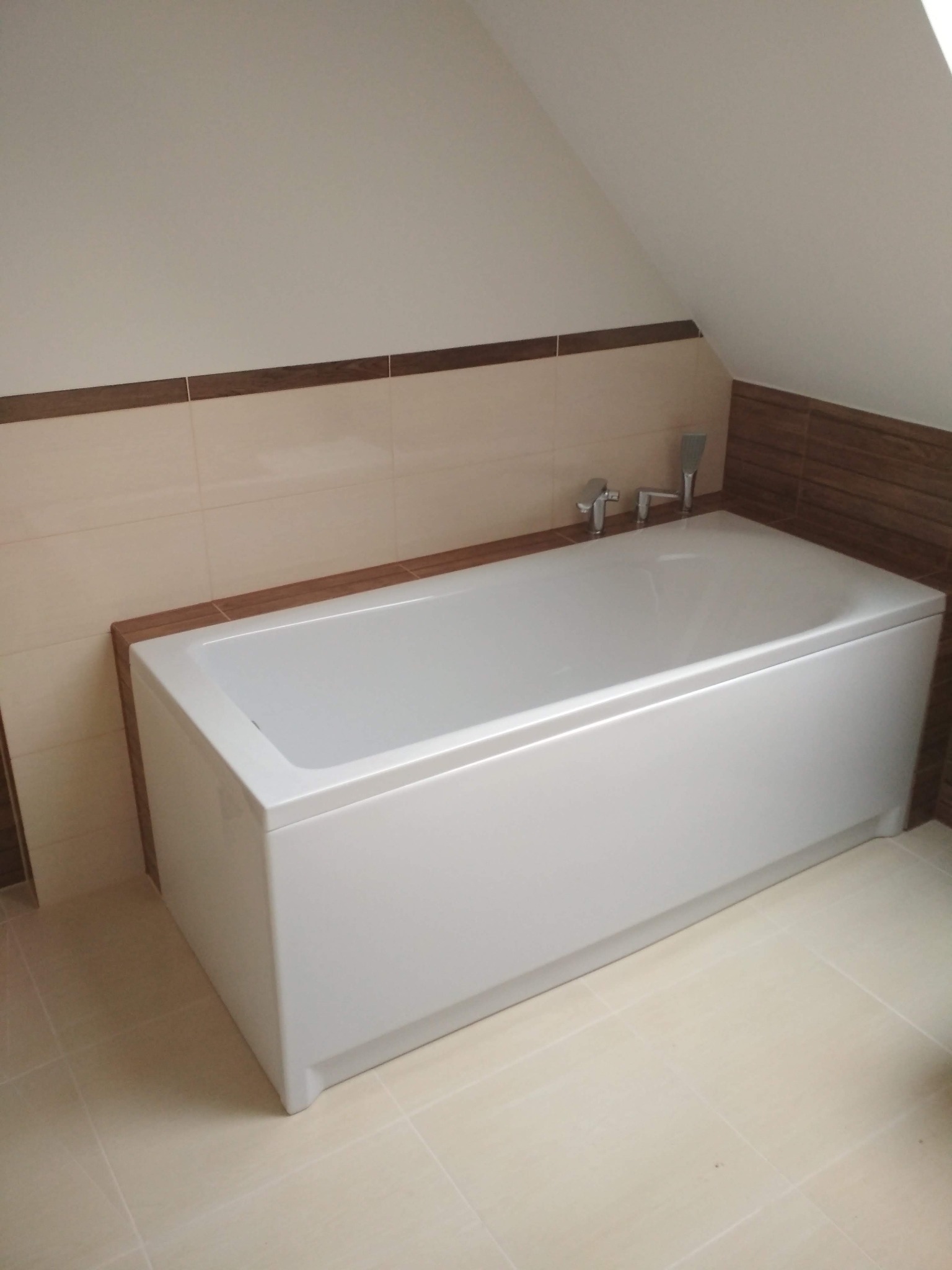 Biała wanna w narożnej łazience na poddaszu, wykończona beżowymi i brązowymi płytkami, widoczna bateria i słuchawka prysznicowa.