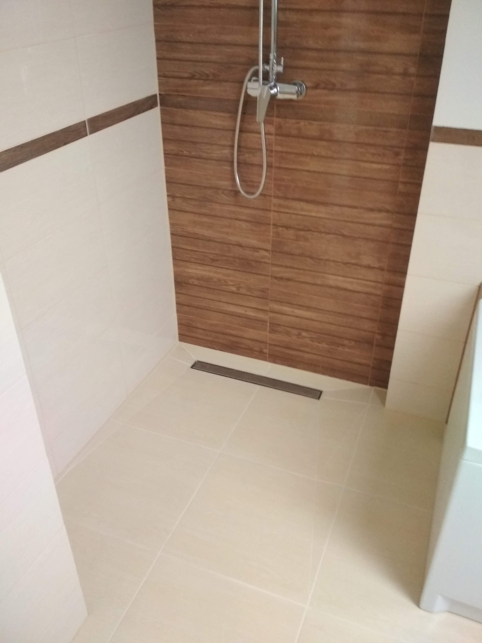 Nowoczesna kabina prysznicowa walk-in z odpływem liniowym, wykończona płytkami imitującymi drewno i beżowymi płytkami z brązowym pasem dekoracyjnym.