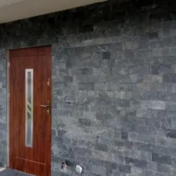 Elewacja z ciemnoszarej, łupanej płytki kamiennej ułożonej na ścianie obok drewnianych drzwi wejściowych z podłużnym, srebrnym przeszkleniem.