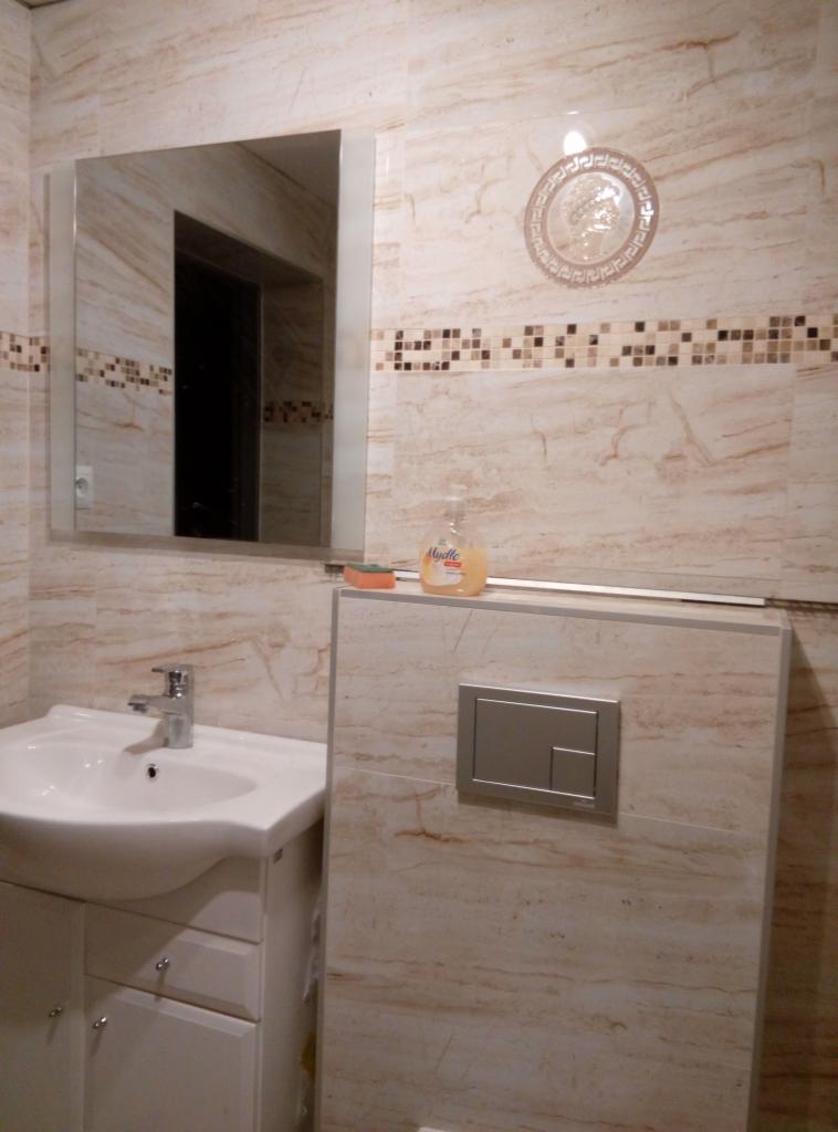 Wykończona łazienka z beżowymi płytkami imitującymi marmur, umywalką z szafką, dużym lustrem i dekoracyjnym medalionem na ścianie.