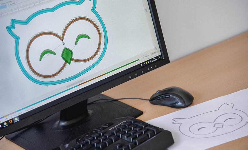 Ekran monitora z projektem graficznym haftu sowy, obok myszka komputerowa i szkic sowy na papierze, klawiatura numeryczna w dolnej części kadru.