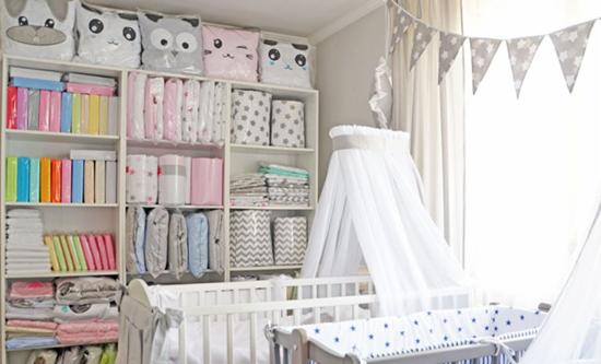 Pokój dziecięcy w jasnych barwach z białym łóżeczkiem pod baldachimem i regałem wypełnionym tekstyliami w pastelowych odcieniach i wzorach, całość dopełniona girlandą z trójkątów.