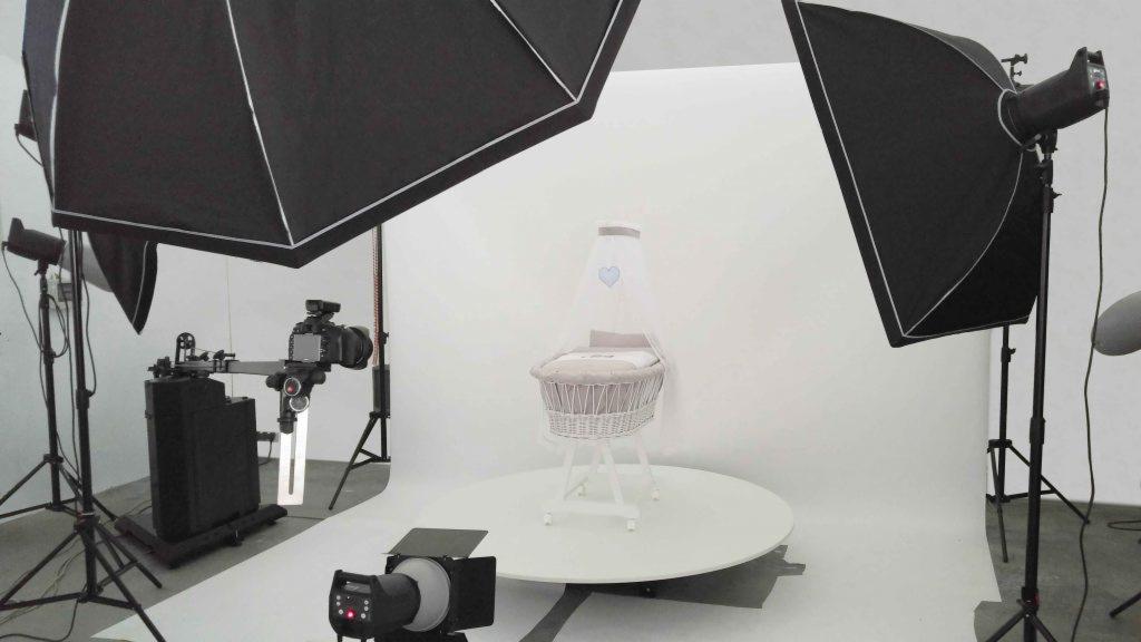 Studio oświetleniowe z profesjonalnym sprzętem fotograficznym otaczającym wiklinowy kosz Mojżesza z białym baldachimem i niebieskim sercem, umieszczony na białym obrotowym podeście.