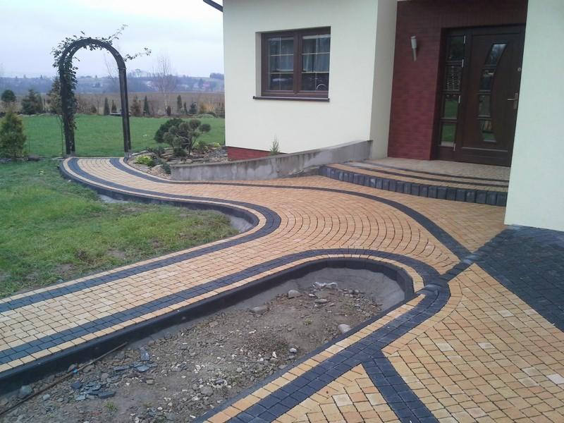 Kręta ścieżka z beżowo-czarnej kostki prowadzi od zielonego trawnika do domu z brązowymi drzwiami. Widoczna brama z roślinami i rabata.