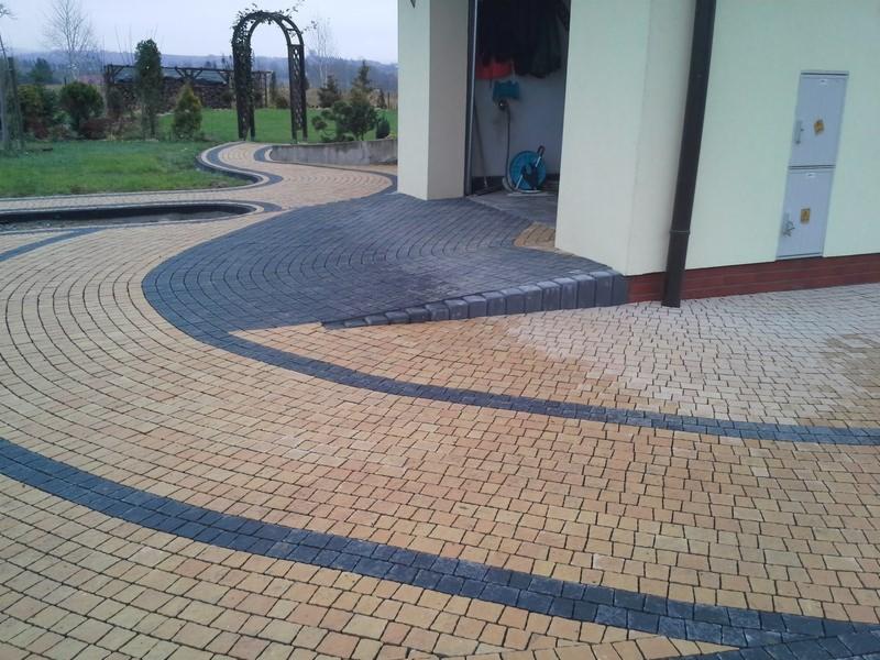 Wzorzysta, łukowata ścieżka z kostki brukowej w odcieniach beżu i grafitu prowadzi do budynku z otwartym garażem i zielonego ogrodu z ozdobną pergolą.