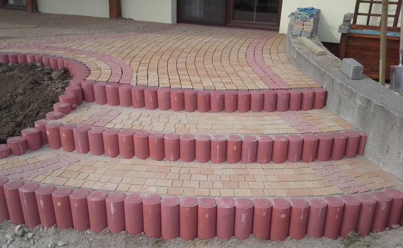 Schody z kostki brukowej w odcieniach beżu i czerwieni, z cylindrycznymi obrzeżami, prowadzące na taras z kostki ułożonej w łukowaty wzór.
