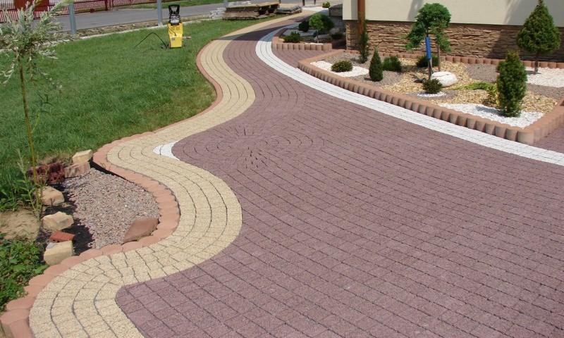 Wzorzysta, brązowa kostka brukowa ułożona w łukowaty wzór, obramowana beżową kostką i białym żwirem. Widoczny trawnik i rabaty z roślinami.