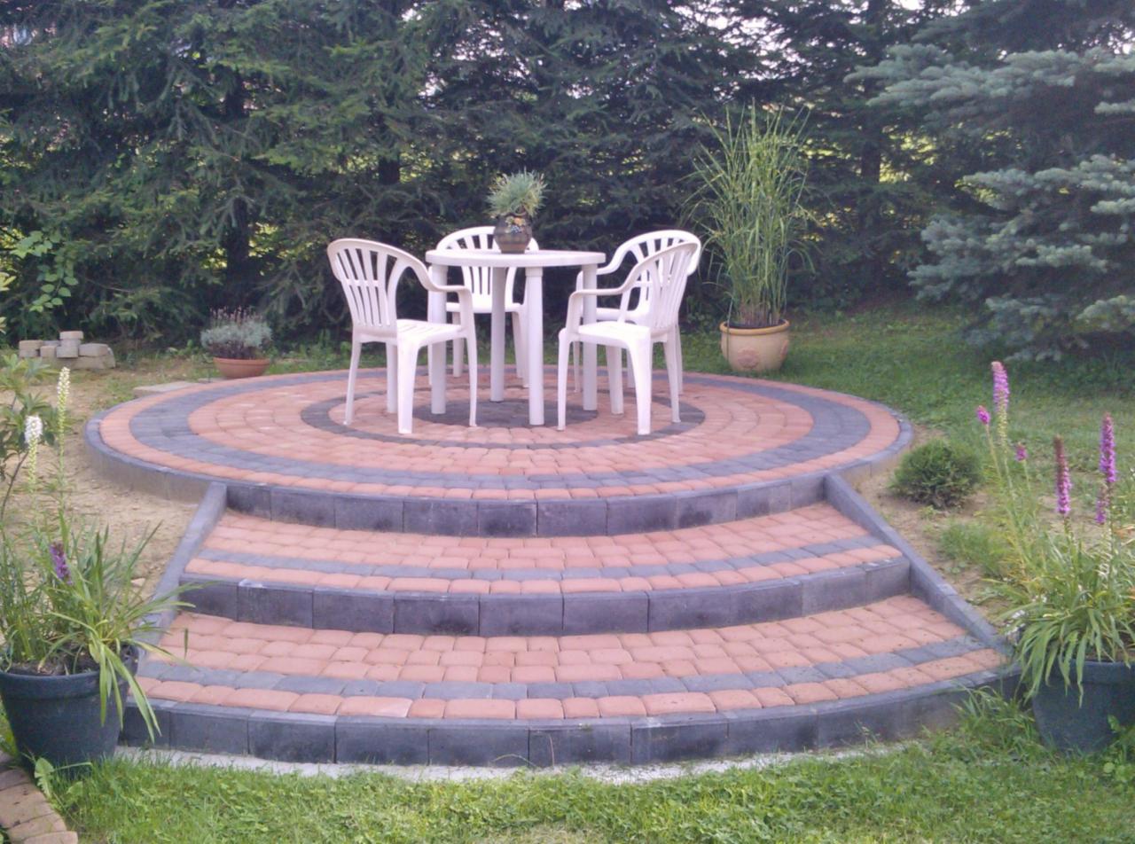 Okrągły taras z kostki brukowej z trzema schodami, otoczony zielenią. Na tarasie biały stół i krzesła ogrodowe.