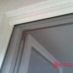 Zamknięta moskitiera w białej ramie, widoczny fragment ściany z fakturą i logo firmy w prawym dolnym rogu.